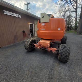 Orange articulated boom lift parked in front of Hibner Sales building with bare trees and utility lines visible
