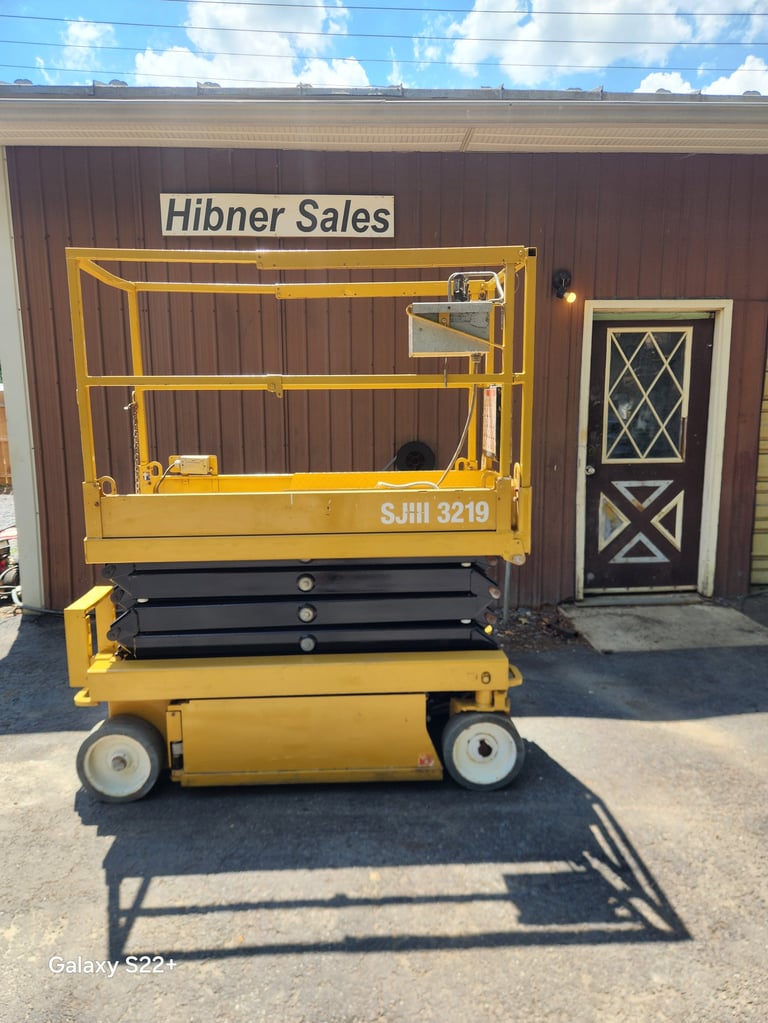 Yellow scissor lift on display at Hibner Sales facility with brown metal building in background