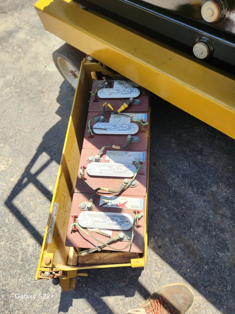 Yellow industrial vehicle with open compartment exposing stacked batteries connected with cables underneath